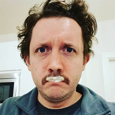 Man scowling humorously to camera, shaving cream half-covering his moustache.