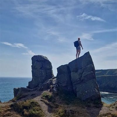 Distant, wide shot of man in hiking gear, standing on ocean cliffs.