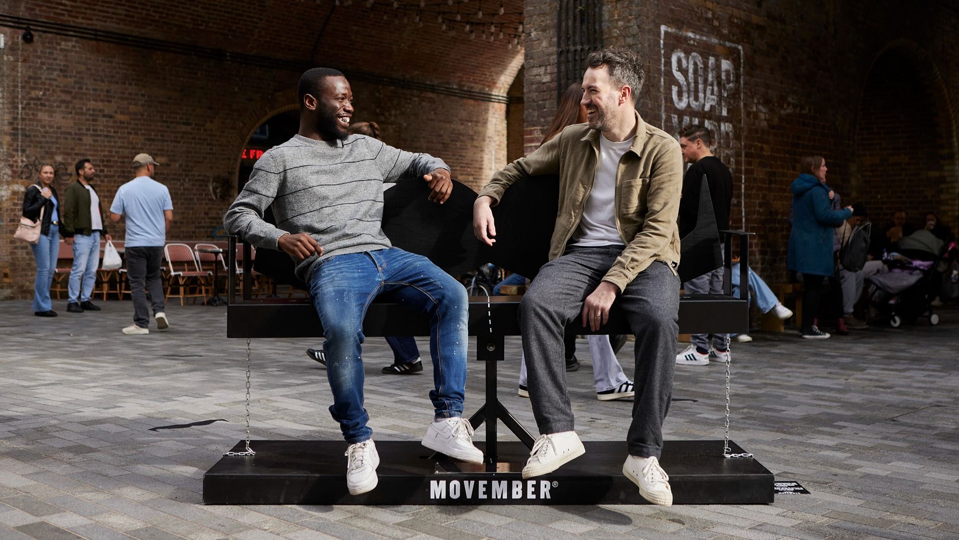 Photo of two smiling men, sitting on a moustache-shaped public bench.